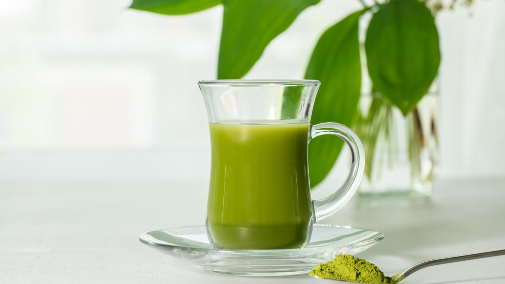 A glass of matcha tea with some powder on a spoon in front of it and a plant in the background